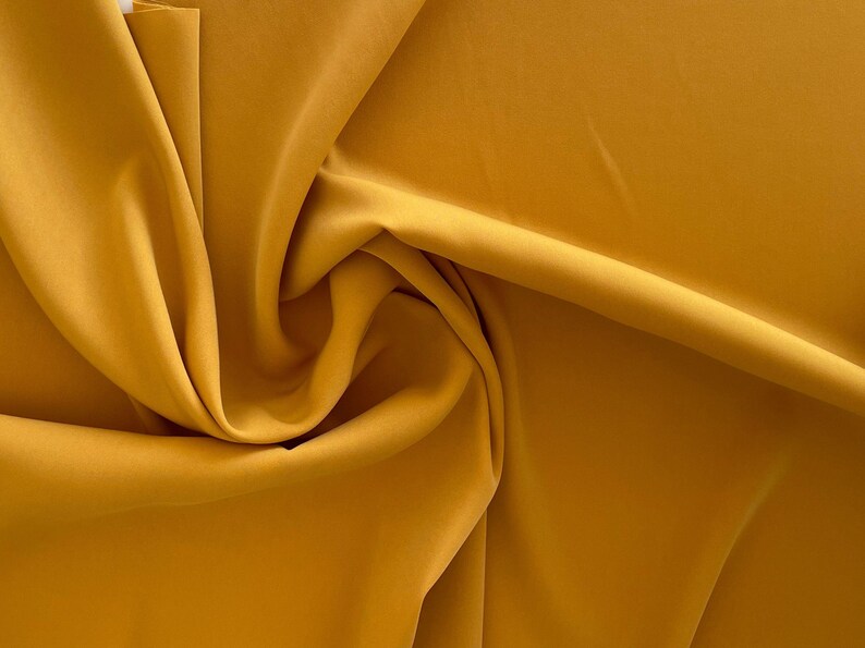 May include: A close-up of a smooth, mustard yellow fabric. The fabric is folded and draped, creating soft folds and curves.