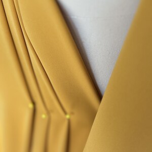May include: Close-up of a mustard yellow fabric with pleats and pins. The fabric is folded and pinned in place, creating a series of vertical lines.