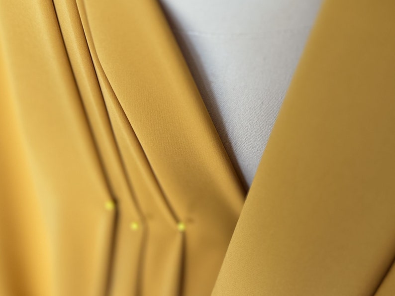 May include: Close-up of a mustard yellow fabric with pleats and pins. The fabric is folded and pinned in place, creating a series of vertical lines.