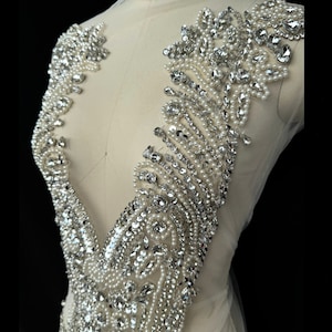 May include: A close-up of a silver and white beaded embellishment. The embellishment is made of small, round beads and rhinestones. It is designed to be sewn onto clothing or other fabric.