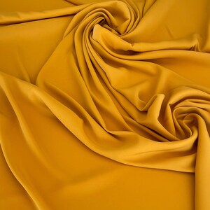 May include: A close-up of a smooth, mustard yellow fabric draped in soft folds. The fabric has a subtle sheen and a luxurious feel.