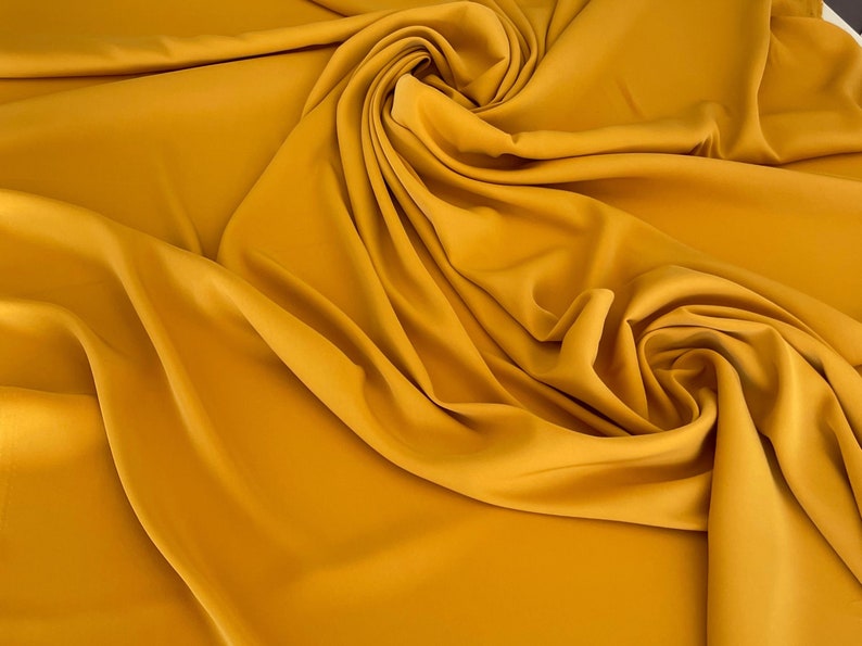 May include: A close-up of a smooth, mustard yellow fabric draped in soft folds. The fabric has a subtle sheen and a luxurious feel.