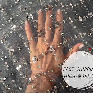 May include: Close-up of a sheer fabric with silver sequins and white beads arranged in a floral pattern. The fabric is delicate and has a soft, flowing texture.