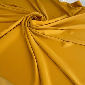 May include: A smooth, mustard yellow fabric draped in soft folds. The fabric has a subtle sheen and a luxurious feel.