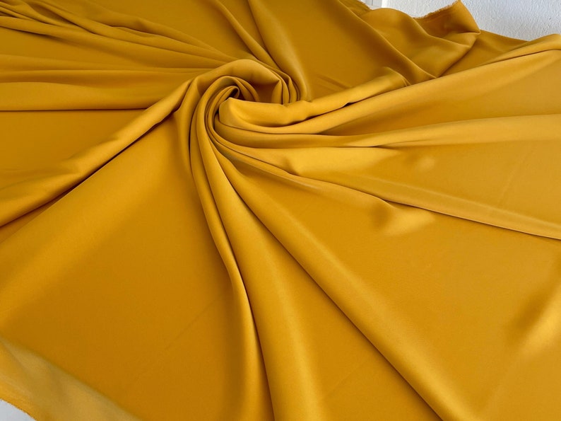 May include: A smooth, mustard yellow fabric draped in soft folds. The fabric has a subtle sheen and a luxurious feel.