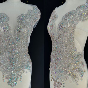 May include: A close-up of a silver and iridescent crystal embellishment for clothing. The embellishment is shaped like a V-neckline and has a floral design. It is sewn onto a white mesh fabric.
