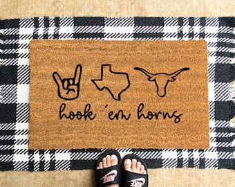 May include: A rectangular doormat with a brown coir center and a black and white plaid border. The center features black line art of a hand gesture, the state of Texas, and a longhorn, with the words "hook 'em horns" below.