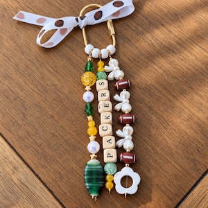 May include: A gold-toned carabiner keychain featuring a white ribbon with brown football patterns. Three beaded strands hang from the carabiner, including green, yellow, and white beads, wooden letter beads spelling PACKERS, and football charms.