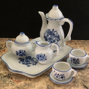 Może przedstawiać: Miniaturowy porcelanowy zestaw do herbaty z białą podstawą i niebieskimi kwiatowymi wzorami. Zestaw zawiera imbryk, dzbanuszek do śmietanki, cukiernicę, dwie filiżanki i spodki, wszystko ułożone na pasującej tacy. Idealny do domku dla lalek.
