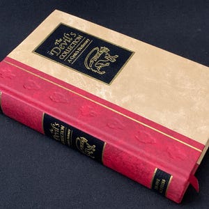 Könnte beinhalten: Ein rot- und cremefarbenes Hardcover-Buch mit dem Titel "The Devil's Collection: A Grimoire Dictionary". Das Buch hat einen goldenen Rücken mit einer schwarzen und goldenen Illustration eines Teufels.
