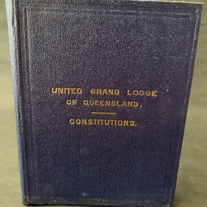 Puede incluir: Un libro azul oscuro con letras doradas que dicen "United Grand Lodge of Queensland. Constitutions."
