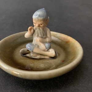 May include: A small ceramic figurine of a leprechaun sitting on a green and brown ceramic dish. The leprechaun is wearing a blue hat and has a white beard.