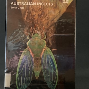 Op de afbeelding: Een boek getiteld "Australian Insects" van John Child. De omslag toont een close-up van een groene cicada met iriserende blauwe vleugels.