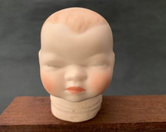 Może przedstawiać: Głowa porcelanowej lalki z zamkniętymi oczami i namalowanym uśmiechem. Głowa lalki jest biała z różowymi policzkami i jasnobrązowym przedziałkiem.