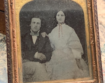 May include: A vintage framed photograph of a couple. The man is wearing a dark suit and the woman is wearing a white dress. The photograph is in a gold-colored frame with ornate details.