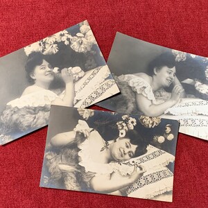 May include: Three vintage black and white photographs of women posing in a studio setting. Each woman is wearing a white dress and is surrounded by flowers. The photographs are printed on thick card stock and have a slightly textured surface.