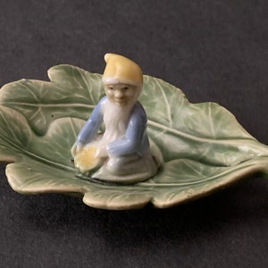May include: A small ceramic figurine of a gnome sitting on a green leaf-shaped dish. The gnome is wearing a yellow hat, blue shirt, and white trousers. The dish is decorated with green leaves.