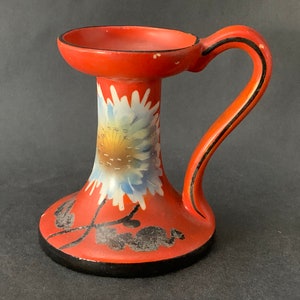 Könnte beinhalten: Roter Kerzenständer aus Keramik mit schwarzem Griff und weißem Blumendesign.