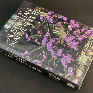 Pode incluir: Um guia de campo para arbustos nativos australianos. A capa do livro apresenta um fundo escuro com um desenho floral de flores roxas e folhas verdes. O título está em texto branco.