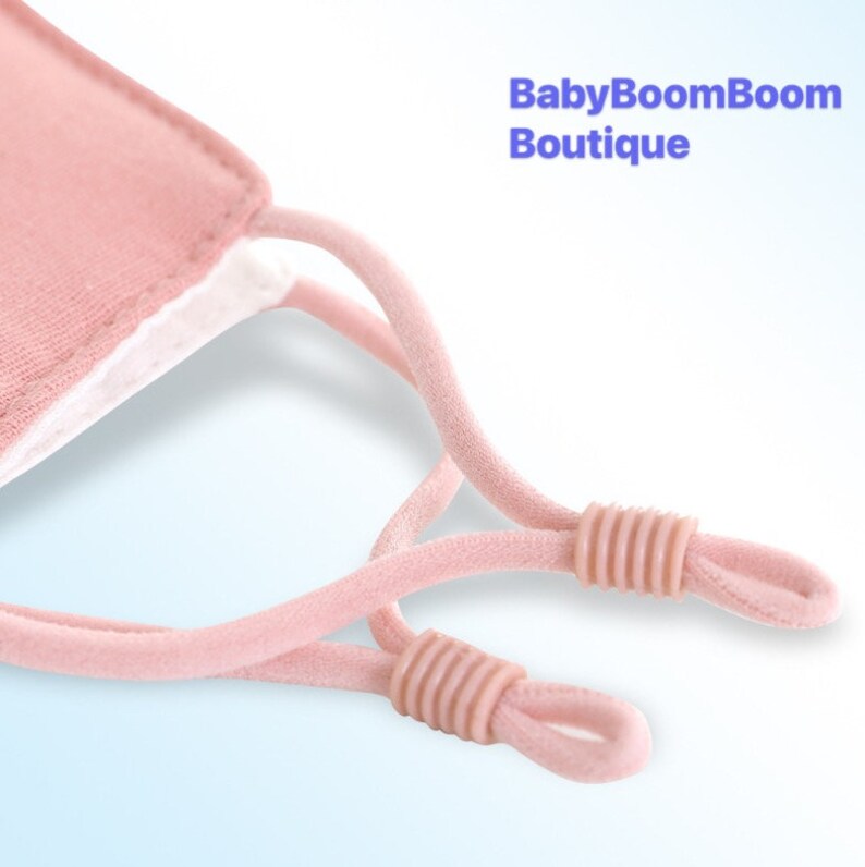 May include: Close-up of the pink elastic ear loops on a pink face mask. The ear loops have adjustable toggles. The mask is made by BabyBoomBoom Boutique.