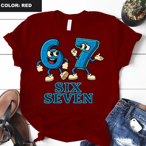 May include: A red t-shirt featuring the numbers six and seven as cartoon characters in blue with white accents. The text "SIX SEVEN" is displayed in blue below the characters. The shirt is a solid red color.