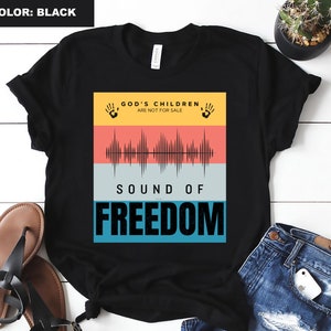 Puede incluir: Camiseta negra con un diseño gráfico que presenta una onda sonora y el texto "God's Children Are Not For Sale" y "Sound of Freedom".