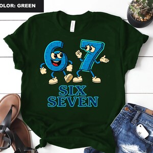 May include: Green t-shirt featuring the numbers six and seven as cartoon characters with arms, legs, and facial expressions. The text "SIX SEVEN" is below the characters in blue. The shirt is on a white surface.