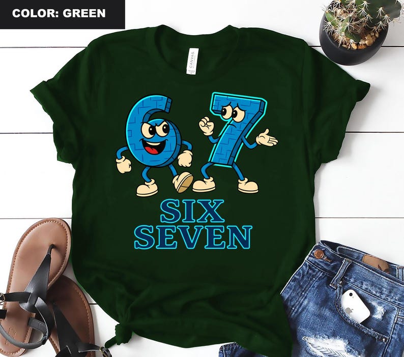 May include: Green t-shirt featuring the numbers six and seven as cartoon characters with arms, legs, and facial expressions. The text "SIX SEVEN" is below the characters in blue. The shirt is on a white surface.