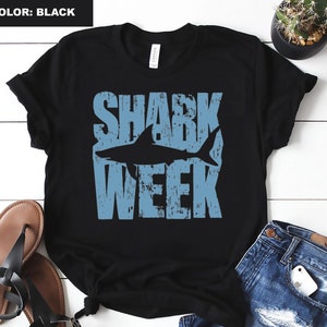 May include: Black t-shirt with a blue graphic that says "Shark Week" with a shark silhouette in the middle of the text.