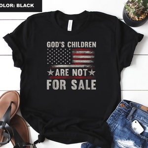 Puede incluir: Camiseta negra con un diseño de bandera estadounidense desgastado y el texto "God's Children Are Not For Sale".