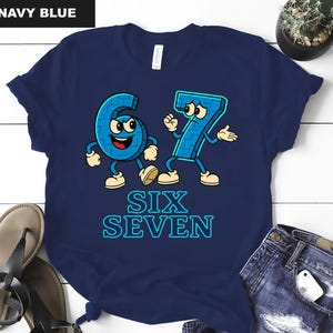 May include: Navy blue t-shirt featuring cartoon numbers six and seven with arms, legs, and facial expressions. The text "SIX SEVEN" is below the numbers in a blue, outlined font. The shirt is a casual, fun design.