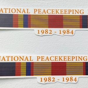 May include: Two stickers featuring a ribbon design with the text "MULTINATIONAL PEACEKEEPING FORCE BEIRUT" and the years "1982 - 1984". The ribbons have stripes of red, yellow, blue, and green.