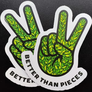 Può includere: Due adesivi bianchi con un segno di pace verde con contorno nero e il testo "Better Than Pieces" in nero.