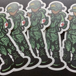 Puede incluir: Cinco pegatinas de camuflaje verde y negro de un personaje de dibujos animados que lleva un sombrero verde y amarillo con la palabra "DOC" en él. El personaje lleva una mochila verde con una cruz roja.