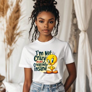 May include: White t-shirt featuring the cartoon character Tweety Bird. The shirt has the text "I'm Not Crazy, I'm Just Creatively Insane" in a dark green and yellow design. The cartoon character is yellow with blue eyes and orange feet.
