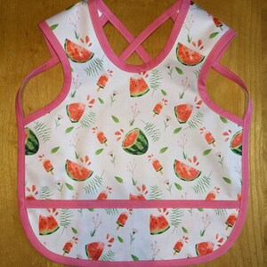 May include: A pink and white bib with a watermelon and popsicle print. The bib has a pocket on the front and a criss-cross strap design.