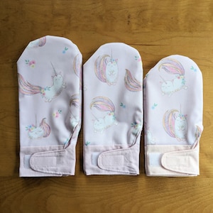 Pode incluir: Três luvas de forno cor-de-rosa com um design de unicórnio. As luvas têm um unicórnio branco com uma crina e uma cauda arco-íris sobre um fundo rosa com pequenos detalhes florais. As luvas têm um fecho de velcro.