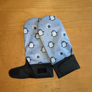 May include: A pair of blue oven mitts with a penguin pattern. The mitts have black straps and velcro closures.