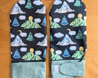 Könnte beinhalten: Ein Paar schwarze Ofenhandschuhe mit einem grünen, blauen und weißen Berg- und Waldmuster. Die Handschuhe haben einen hellblauen Bund und einen Riemen an einem Handschuh.