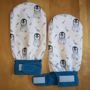 Puede incluir: Dos manoplas de horno azul turquesa con fondo blanco y un patrón repetido de pingüinos con pajaritas azules. Las manoplas tienen un cierre de velcro.