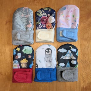 May include: Six pairs of baby mittens with different patterns. The mittens are made of soft fabric and have a fleece lining. The patterns include flamingos, flowers, a unicorn, owls, a penguin, and a mountain.