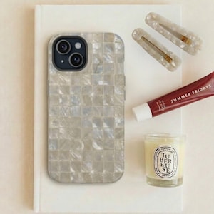May include: An assortment of beauty and lifestyle items arranged on a white surface. Includes a pearlescent phone case, two hair clips, a tube of Summer Fridays lip balm, a Diptyque candle, and a white notebook.