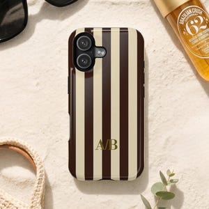 Op de afbeelding: Een smartphonehoesje met een verticaal gestreept patroon in donkerbruin en crèmekleur. In het midden van het hoesje staan de goudkleurige initialen A/B in een schreeflettertype. Het hoesje heeft een uitsparing voor een dubbele camera linksboven.