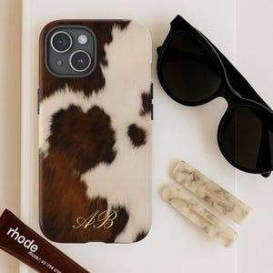 Puede incluir: Una funda de teléfono con estampado de piel de vaca marrón y blanco con iniciales doradas, gafas de sol negras, dos pinzas para el pelo de mármol y un tubo de pintalabios marrón. Los artículos están colocados sobre una superficie blanca.
