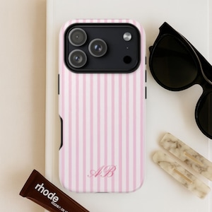 Op de afbeelding: Een roze en witte gestreepte telefoonhoes met de initialen "AB" in een sierlijk schrift. De hoes ligt naast een zwarte zonnebril, een bruine tube "rhode" lippenbalsem en twee marmeren haarclips. De hoes is ontworpen voor een moderne smartphone.