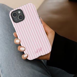 Op de afbeelding: Een roze en wit gestreept telefoonhoesje met de initialen "AB" in een scriptlettertype. De hoes wordt in een hand gehouden en toont de camera van de telefoon en de zwarte rand.