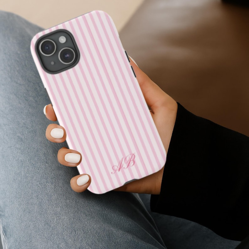 Op de afbeelding: Een roze en wit gestreept telefoonhoesje met de initialen "AB" in een scriptlettertype. De hoes wordt in een hand gehouden en toont de camera van de telefoon en de zwarte rand.