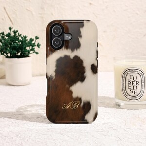 Puede incluir: Una funda rectangular para smartphone con un estampado de piel de vaca en color marrón y blanco. La funda tiene un recorte para cámara de doble lente en la parte superior izquierda y las iniciales AB en cursiva dorada en el centro.