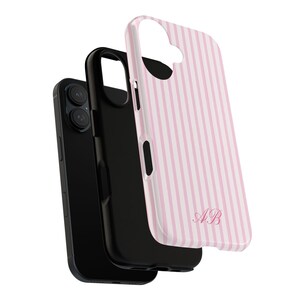 Op de afbeelding: Een zwarte smartphone met een zwarte hoes en een roze en wit gestreepte telefoonhoes. De roze hoes heeft de initialen "AB" in een cursief lettertype. De telefoon en hoesjes worden tegen een witte achtergrond weergegeven.