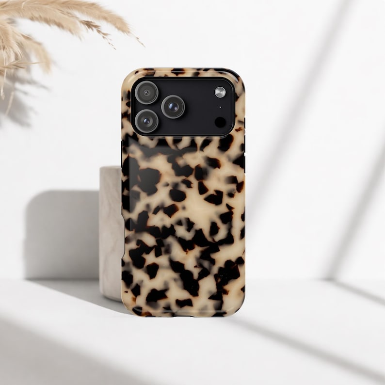 May include: A tortoiseshell-style phone case with a glossy finish. The case has a black camera module with three lenses. The case is tan and brown with a speckled design. The phone case is on a white surface.